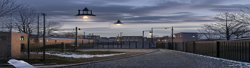 Panoramic view of a modern Irish state detention-campus exterior at dusk, low-rise brown-brick and grey-clad institutional buildings set behind a tall steel perimeter fence with overhead lamps just switching on, a gravel path through a gated courtyard, winter trees in the background, muted industrial palette