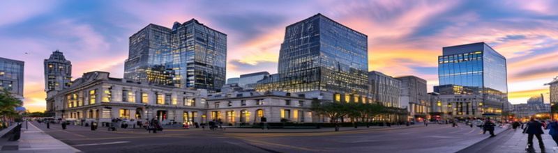 Panoramic view of a modern London financial district skyline at early morning, Bank of England's classical stone facade in the foreground, glass towers of the City rising behind in soft haze, lights still on in upper office floors, empty cobbled street, pale yellow sunrise light