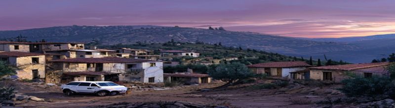 Panoramic view across a southern Lebanese hillside village at dusk, partially destroyed concrete houses with rebar exposed, a white UN vehicle parked on a dirt road below an olive grove, distant ridge of cedar-covered mountains, soft mauve evening light, no people no faces no hands