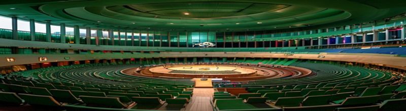 Panoramic view of the United Nations General Assembly hall in New York from a high gallery, the curved tiered green seating arrangement and the wide rostrum platform empty before the session begins, the iconic gold UN emblem and the long row of national flagpoles outside visible through tall windows, soft directional lighting, no people no faces no hands