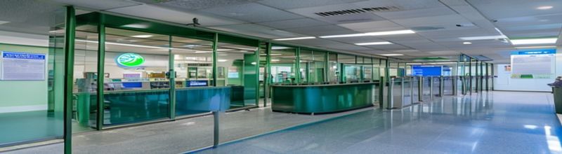 Panoramic view of an Intreo public employment office reception in Dublin at opening time, a glass entrance with the green Intreo logo decal, an empty queue rail with retractable belts, a digital ticket-number display showing a single illuminated number, a noticeboard with printed leaflets, polished blue-grey floor, no people no faces no hands