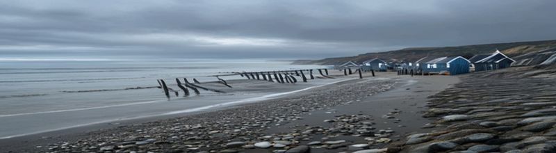 Panoramic coastal view at Whitstable Kent at low tide, a wide flat shingle beach under grey morning sky, a row of weathered black-painted fishermens huts on the beach line, a long wooden groyne running into the calm sea, a faint plume of foam where a brook outfall meets the tideline, distant chalky cliffs in the haze, no people no faces no hands