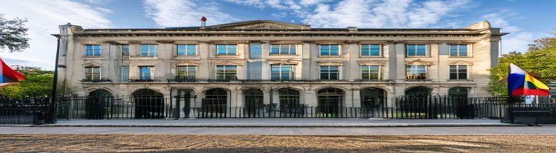 Panoramic view of the front façade of Leinster House in Dublin in afternoon light, classical pediment and tall multi-paned windows above iron railings, the gravel forecourt empty, a tricolour and EU flag mounted on poles to one side, no people no faces no hands