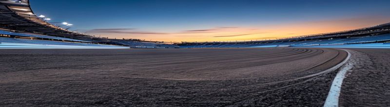 Panoramic view of an empty 400-metre athletics running track at dawn, the lanes painted bright red, white lane lines stretching away into a soft morning haze, a stand of trees on the far side and a single bank of grey grandstand seating to the left, no people no faces no hands