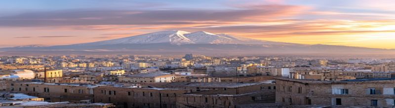 Panoramic morning view of Yerevan with the snow-capped twin peaks of Mount Ararat rising over the southern horizon, terracotta-stoned Soviet-era buildings in the foreground catching pale gold sunrise light, a wide pedestrian boulevard running into the distance, a single Armenian tricolour and an EU flag on tall flagpoles outside a government compound, no people no faces no hands