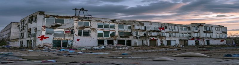 Panoramic exterior view of a damaged hospital building at dusk, broken windows, sandbagged entrance, faded red cross emblem on white wall, scattered medical supplies on the ground, overcast sky, documentary photography style, muted colour palette, no people no faces no hands