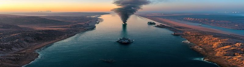 Panoramic aerial view at dawn of a narrow ocean strait between rocky desert coastlines with grey naval destroyers forming a line on calm water and a column of black smoke rising from an oil terminal complex on the far shore, cargo tankers anchored in the distance, no people no faces no hands