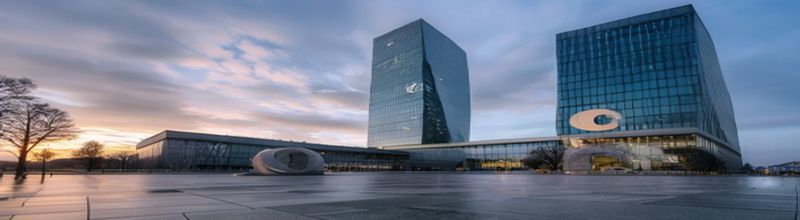 Panoramic exterior of the European Central Bank Eurotower in Frankfurt at dawn, glass facade reflecting sky, euro symbol sculpture on stone plinth in foreground, calm overcast morning, no people no faces no hands