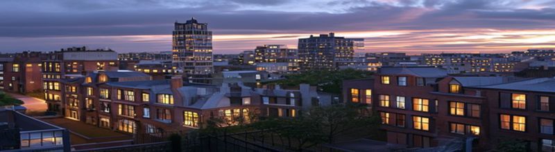 Panoramic aerial view of a mixed British townscape at dusk, rows of brick Victorian terraces in the foreground, a 1960s concrete office tower mid-frame, modern glass-clad commercial buildings in the distance, warm window lights starting to come on, documentary realism, no people no faces no hands