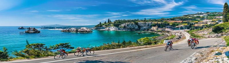 Panoramic wide-angle view of a Giro d'Italia race convoy on a Mediterranean coastal road in soft late-morning light, a small group of cyclists in colourful kit on a curve of pale asphalt high above a brilliant blue-green sea, pale limestone cliffs and tufts of green scrub at the roadside, an indistinct distant headland under a clear bright sky, sharp natural detail on the road surface and barriers, no people no faces no hands