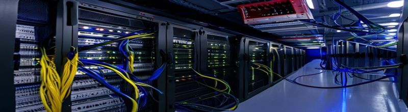 Panoramic view of a darkened server room aisle with two rows of black networking racks, blinking blue and amber LED status lights on a Cisco SD-WAN controller's front panel, neat bundles of yellow and blue Ethernet cabling running along overhead trays, a wall-mounted digital clock reading 17:31, a small red emergency stop button on the cabinet, no people no faces no hands