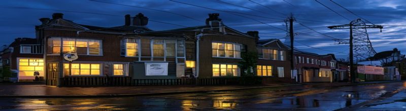Panoramic view of a row of Irish two-storey terraced houses on a Dublin street at twilight with warm yellow kitchen window light spilling onto the wet pavement, an ESB electricity meter cabinet mounted on a side wall, overhead suburban power lines silhouetted against a deep indigo sky, a printed household electricity bill envelope on a doorstep, no people no faces no hands