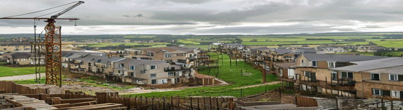 Panoramic view of a new housing estate under construction on the edge of an Irish town, rows of part-built two-storey houses with bare blockwork and scaffolding, a tower crane, stacks of timber and roof tiles, green fields beyond, soft overcast light, no people no faces no hands
